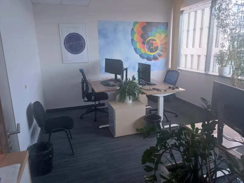 Phone photo of a small open office: two desks with monitors, a hot-air balloon poster, plants and a window wall, with spare chairs and bins in the way